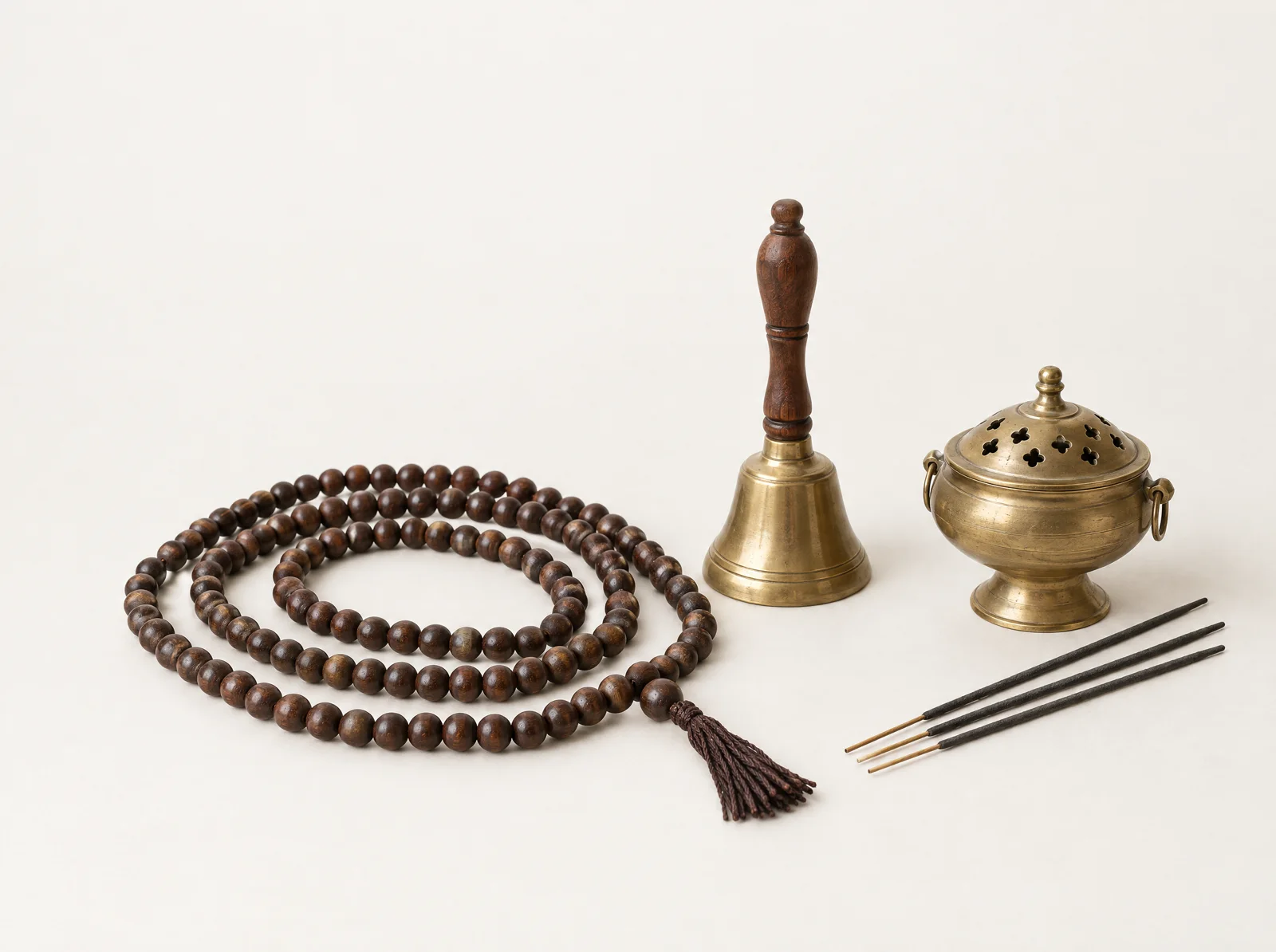 Mala beads, a small brass bell, a censer and three incense sticks on a bone-white background